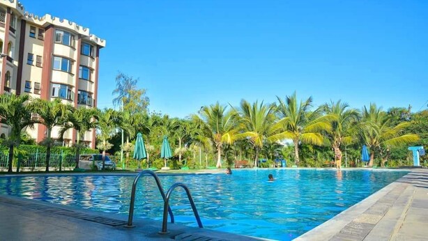 A swimming pool with a clear blue sky, surrounded by palm trees and a multi-story building with balconies. - Blodriff Apartments (Mombasa)