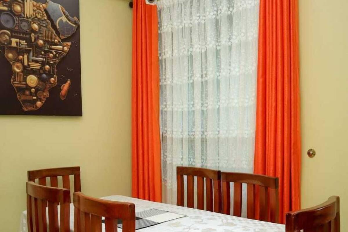 A dining area with a table set for four, surrounded by wooden chairs, a black curtain rod with orange curtains, and a wall-mounted artwork.