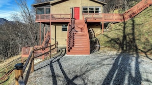 Une cabane à deux étages avec une porte rouge et un balcon, entourée d’arbres et de collines.