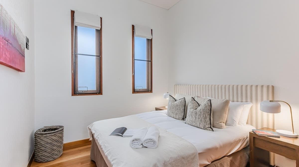 A bedroom with a bed, two windows, a bedside table with a lamp, and a waste basket.