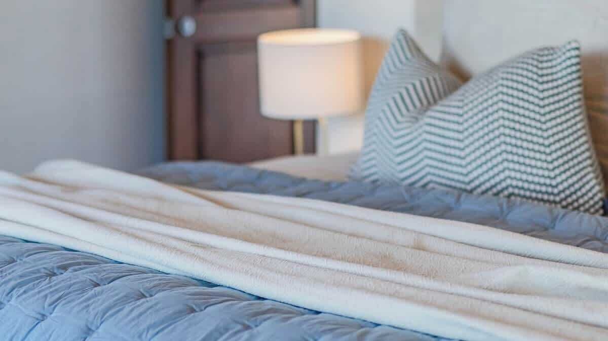 A bed with a quilted cover and pillows, a bedside lamp, and a wooden door in the background.