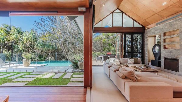 A modern living room with a large sofa, a fireplace, and a view of a swimming pool and garden.