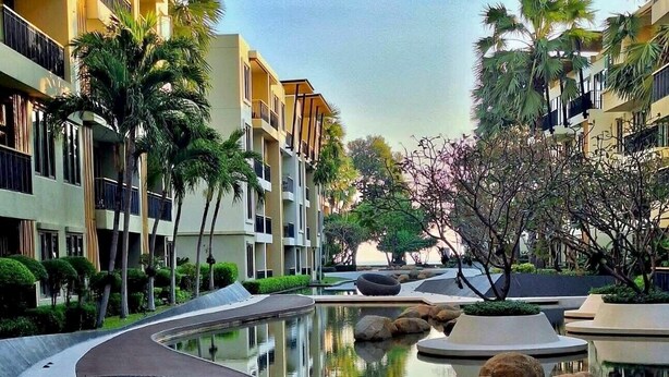 A modern hotel with a pool area, featuring a curved pathway, palm trees, and a reflection pool.