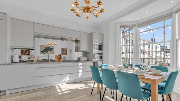 A modern kitchen with white cabinetry, a central island, and a dining area with teal chairs and a wooden table. - City Chic Townhouse | By My Getaways (Brighton)