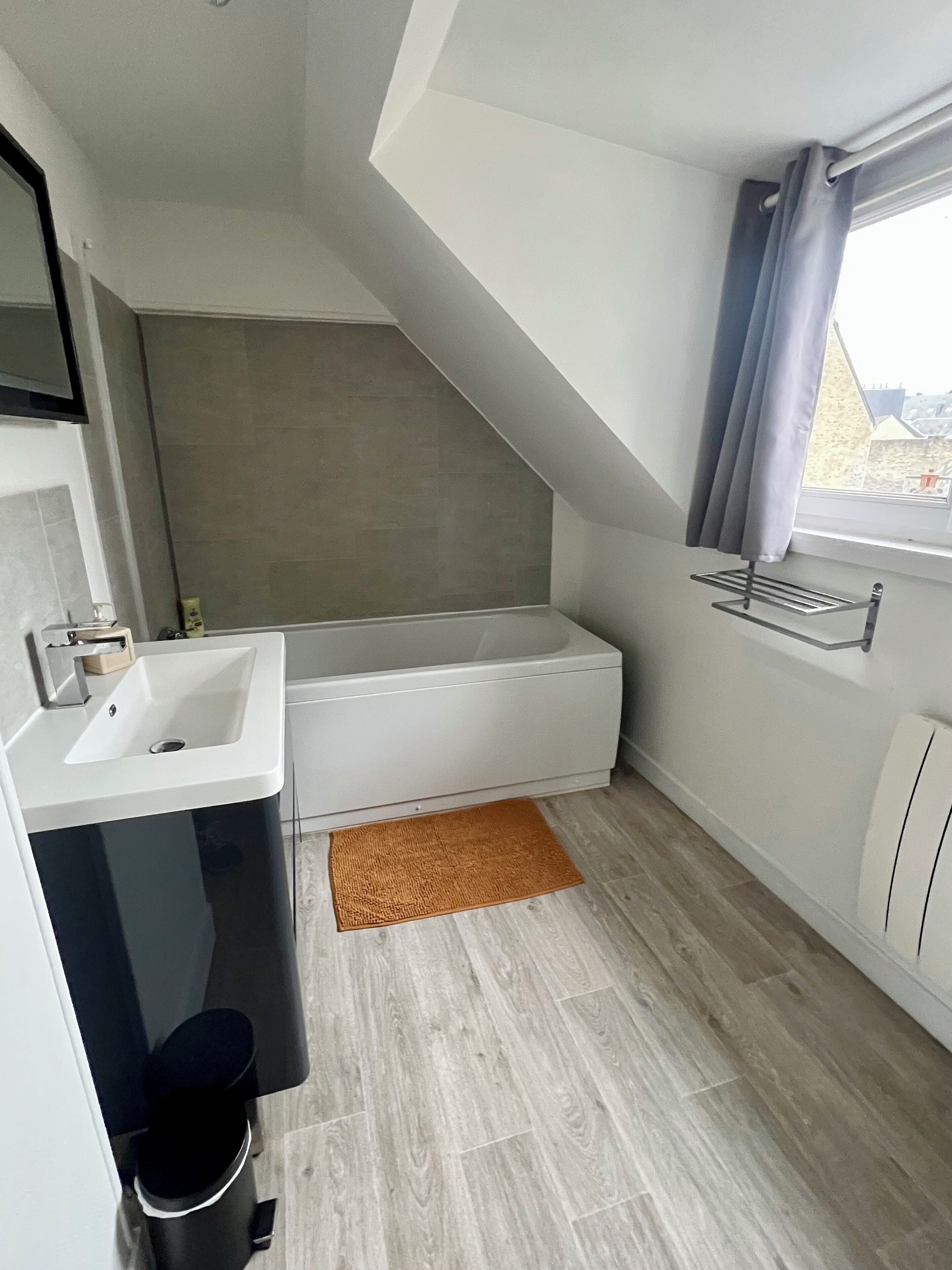 A modern bathroom with a bathtub, a sink, and a toilet. There is a window with curtains, a towel rack, and a wooden floor.