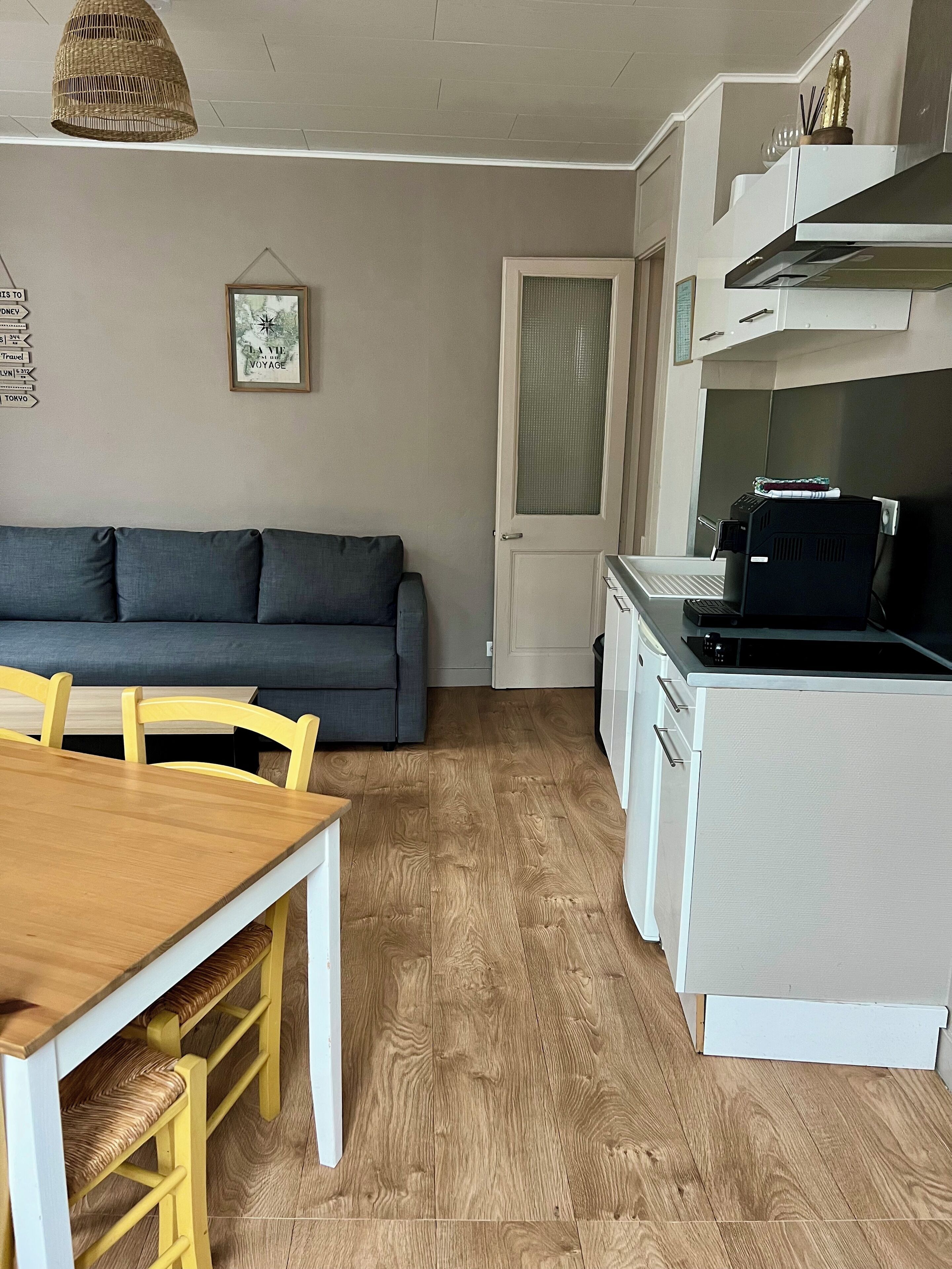 A compact living space with a kitchenette, a dining area with a wooden table and yellow chairs, a gray sofa, and a framed travel poster on the wall.