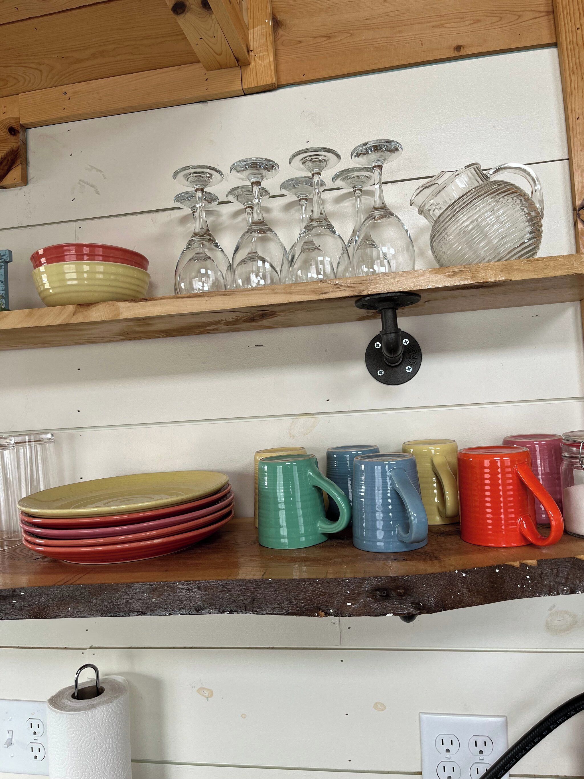 A kitchen shelf with stacked plates, glassware, and colorful mugs.