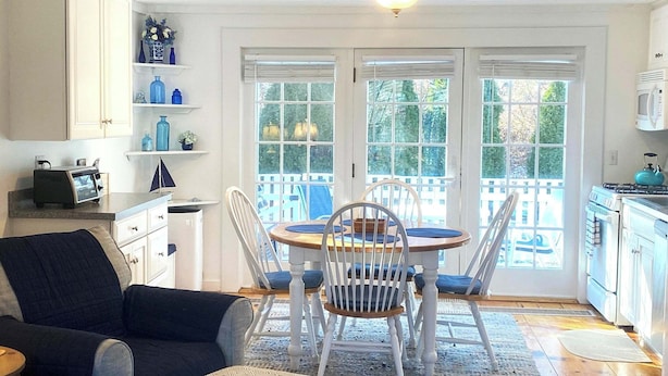 A cozy living room with a dark-colored sofa, a dining table with white chairs, and a kitchen area with white cabinets and a stove. - Maushop Village, 2-bedroom, 1.5 baths, Sleeps 4, A/C, patios & private beach (Mashpee)