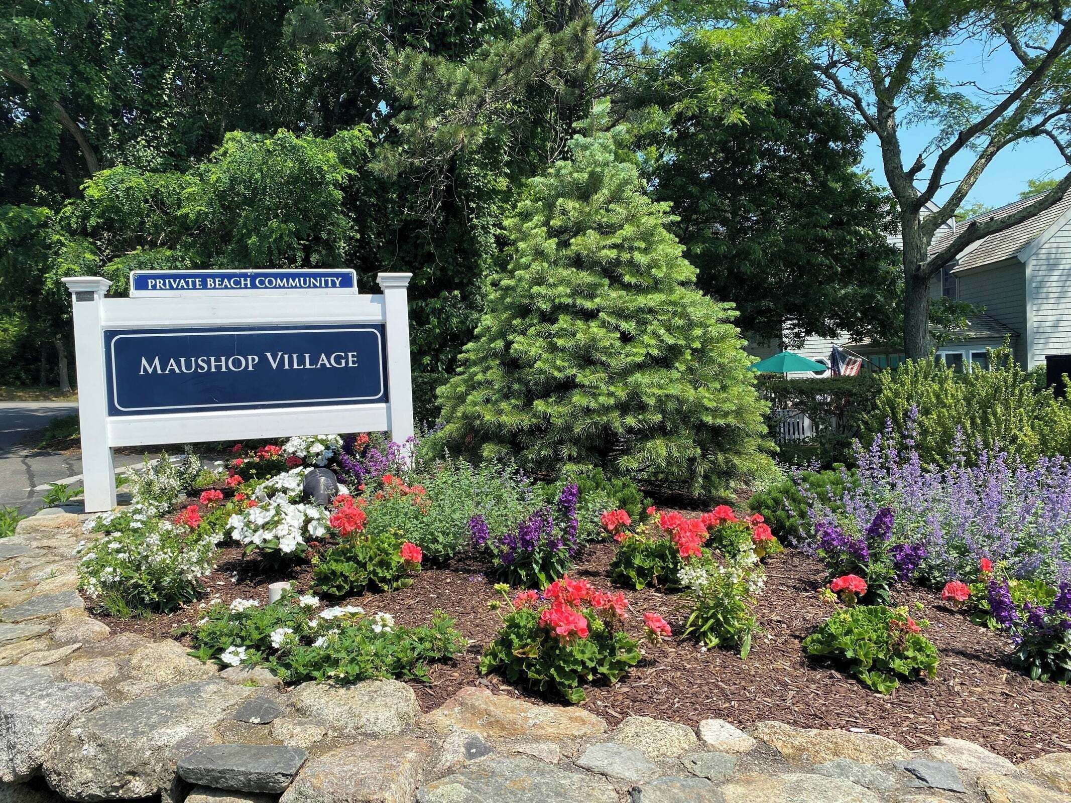 Une enseigne indiquant l’entrée du village de Maushop, avec un jardin rempli de fleurs aux couleurs vives et un pin.