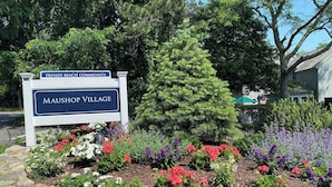 A sign for Maushop Village with a garden of colorful flowers and a pine tree. - Maushop Village, 2-bedroom, 1.5 baths, Sleeps 4, A/C, patios & private beach (Mashpee)