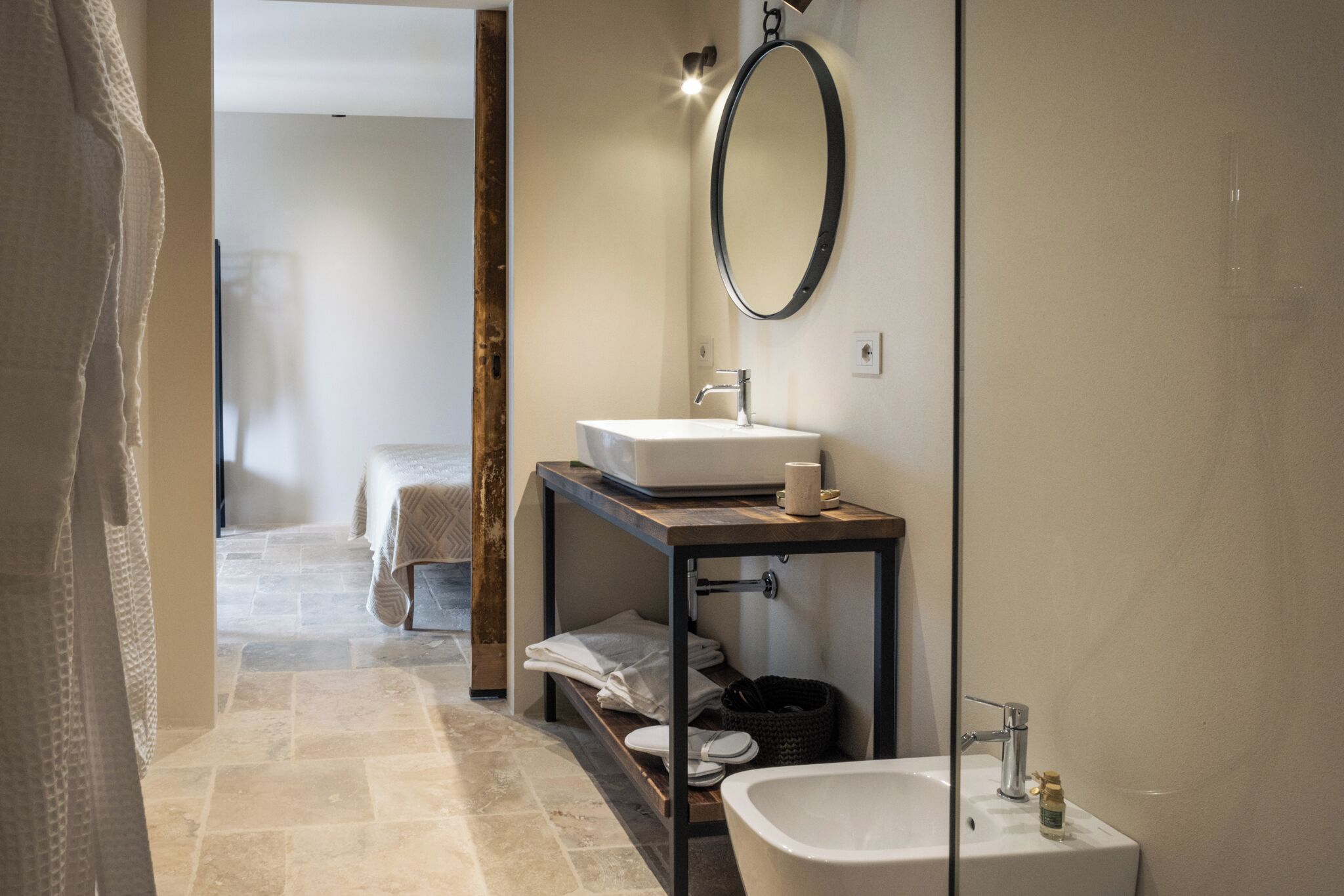 Un baño moderno con un lavamanos blanco, un espejo y una bañera. Hay un estante de madera con toallas y un cubo de basura pequeño.