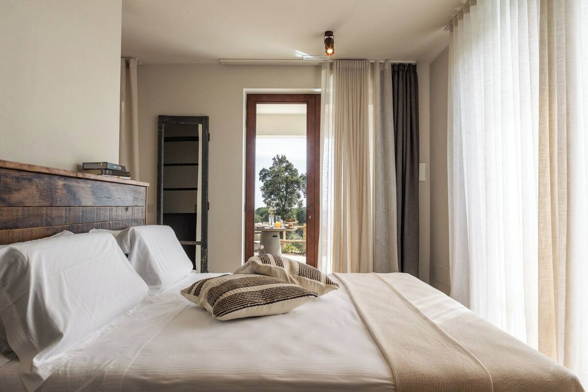 A bedroom with a bed, pillows, a wooden headboard, a wardrobe, and a view through an open door.