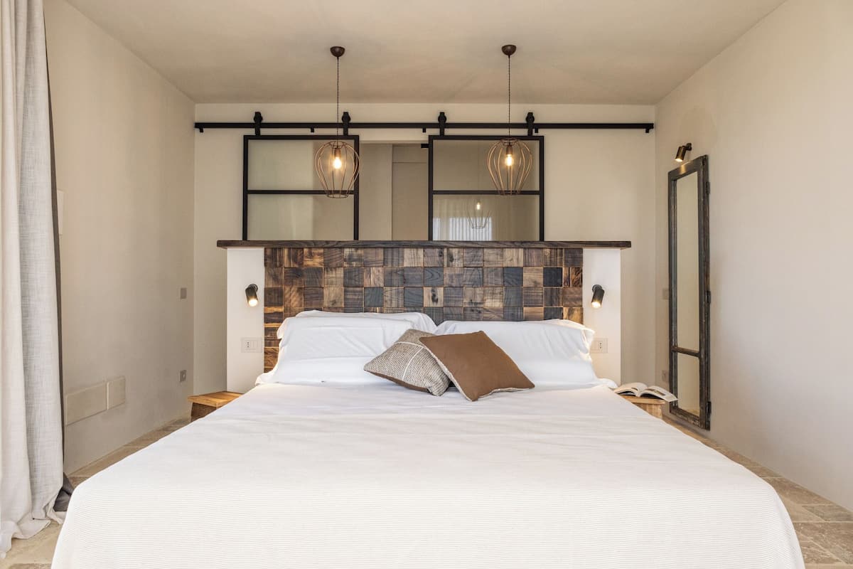 A neatly made bed with white linens and a brown pillow, a wooden headboard, and two pendant lights above.