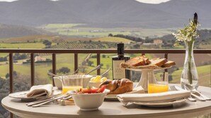 A breakfast table with a scenic view of rolling hills, a vase with flowers, and a bottle of olive oil.