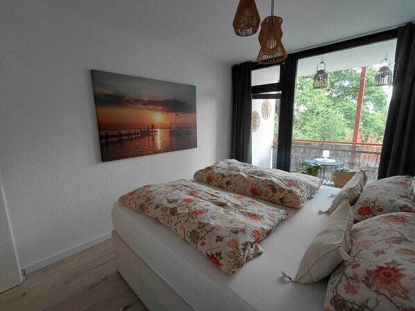 A bedroom with a bed, a painting of a sunset, and a view of greenery through the window.