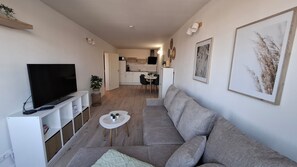 Una sala moderna con un sofá gris, un televisor de pantalla plana, una pequeña mesa redonda con una planta y una cocina al fondo.