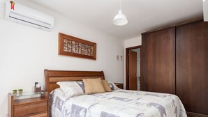 Un dormitorio con cama, cabecero de madera, mesitas de noche, mesita auxiliar, aire acondicionado instalado en la pared, lámpara colgante, armario de madera y un adorno en la pared.