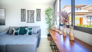 Ein modernes Wohnzimmer mit grauem Sofa, abstrakter Wandkunst, einem Holztisch mit Dekoration und Blick auf ein Gebäude durch ein großes Fenster.