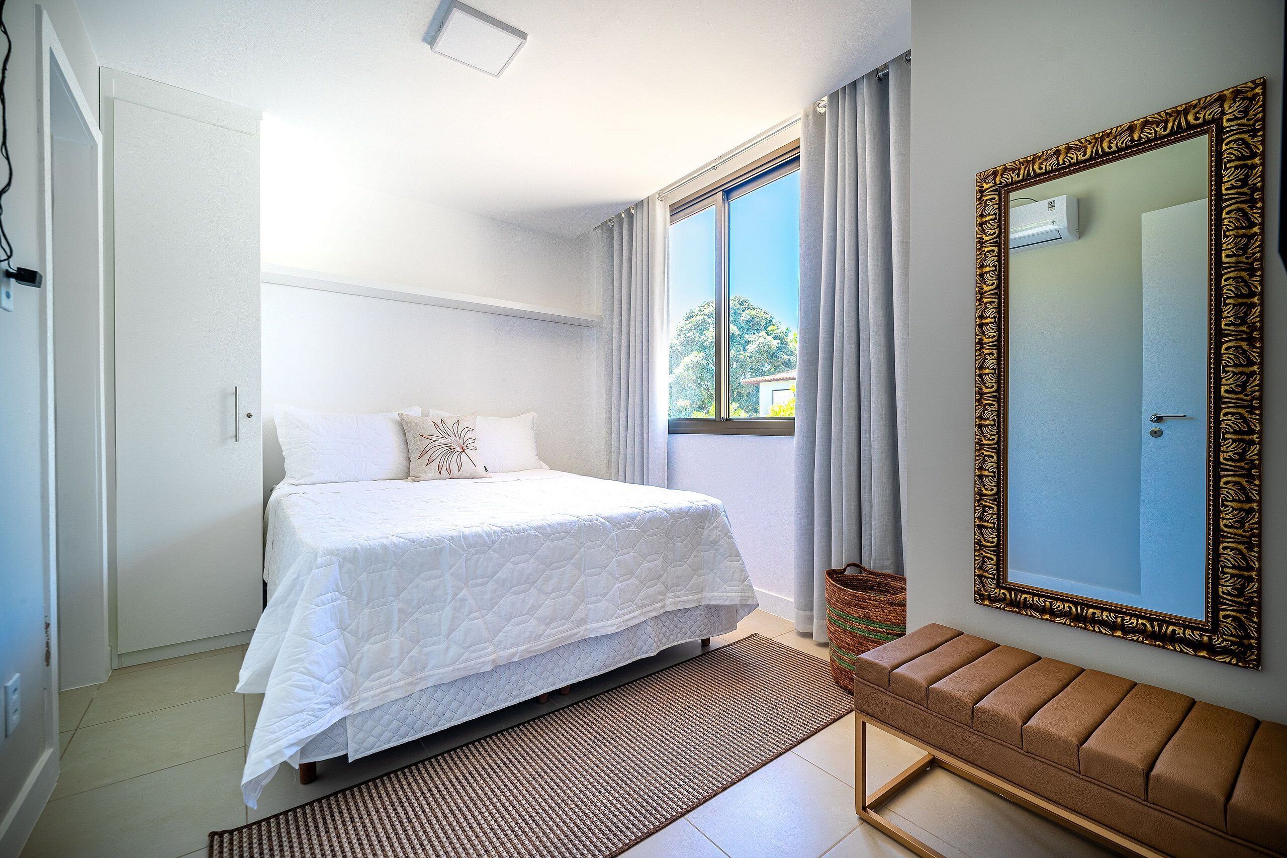 A bedroom with a bed, a bench, a large mirror, and a window with curtains.