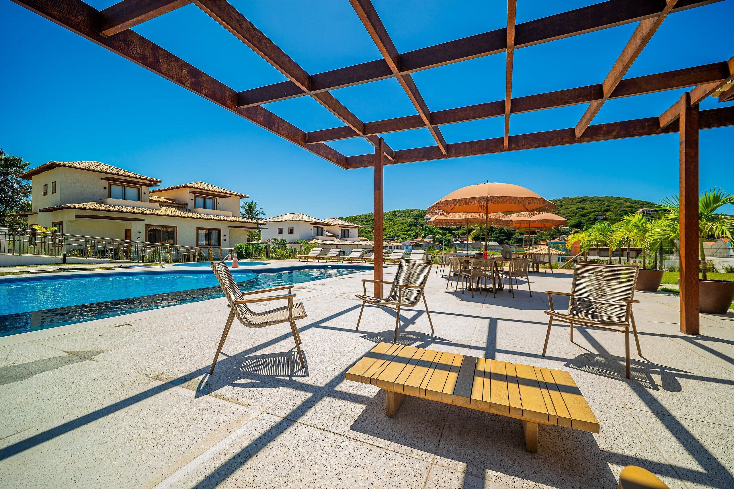 Uma área ao lado da piscina com espreguiçadeiras, uma mesa de madeira e uma estrutura de pérgola.
