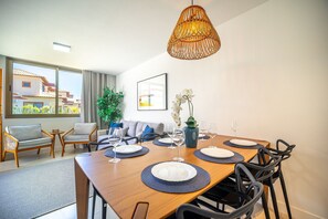 Un espace de salle à manger moderne, avec une table en bois dressée pour six personnes, entourée de chaises noires. Un luminaire suspendu en fibres tressées est accroché au plafond, et une grande fenêtre offre une vue sur un bâtiment à l’extérieur.