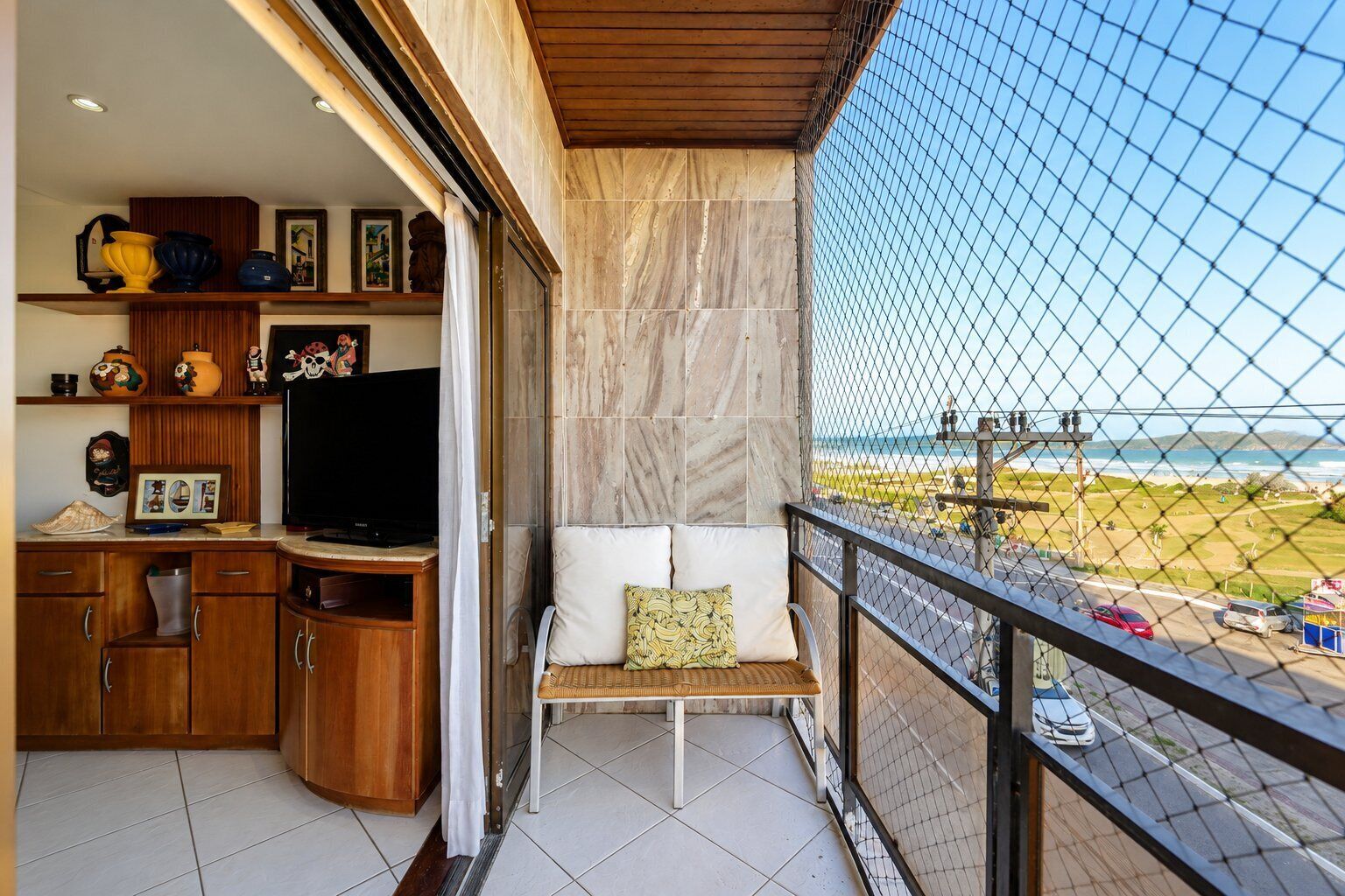 A balcony with a view of the beach, a seating area with cushions, and a TV on a wooden cabinet.