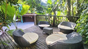 Une terrasse en bois, agrémentée de meubles en osier et d’une petite table, le tout entouré d’une végétation luxuriante.