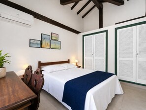Une chambre d’hôtel comprenant un lit, un bureau, une chaise, une plante en pot et un mur orné de trois tableaux encadrés.