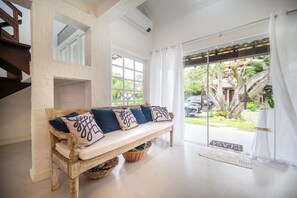 A wooden bench with cushions, a white wall, and a glass door leading to an outdoor area with greenery.