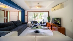 A modern living room with a sectional sofa, two black chairs, a glass coffee table, a TV, and a view of greenery outside.