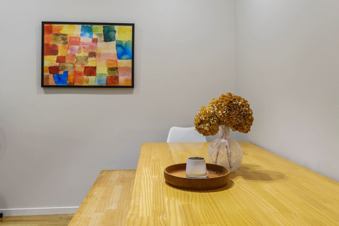 Ein Holztisch mit einer Vase getrockneter Blumen und einer kleinen Kerze. An der Wand hängt ein farbenfrohes, abstraktes Gemälde.