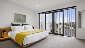 A bedroom with a bed, a bedside table, a lamp, a painting, and a large window with a view of buildings. - Modern Orewa Delight w Parking just Steps to Beach (Auckland Hibiscus Coast)