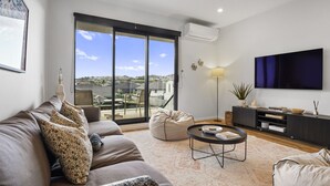 A modern living room with a sofa, a coffee table, a TV, and a view of a cityscape through large windows. - Modern Orewa Delight w Parking just Steps to Beach (Auckland Hibiscus Coast)