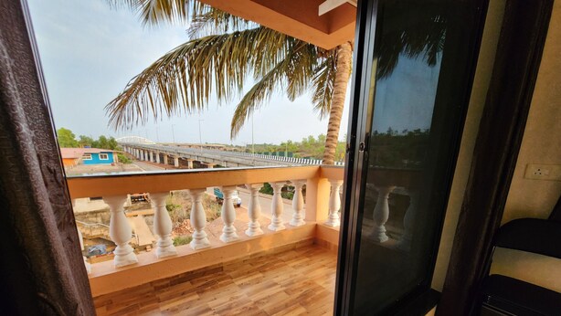 A balcony with wooden flooring and white balustrades, overlooking a road with vehicles and a bridge in the distance. - Sai Sharad Hotel  (Pernem)