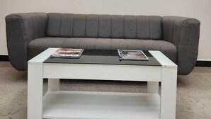 A grey sofa with a white coffee table in front, two magazines on the table, and a framed artwork with a light fixture above. - Sai Sharad Hotel  (Pernem)