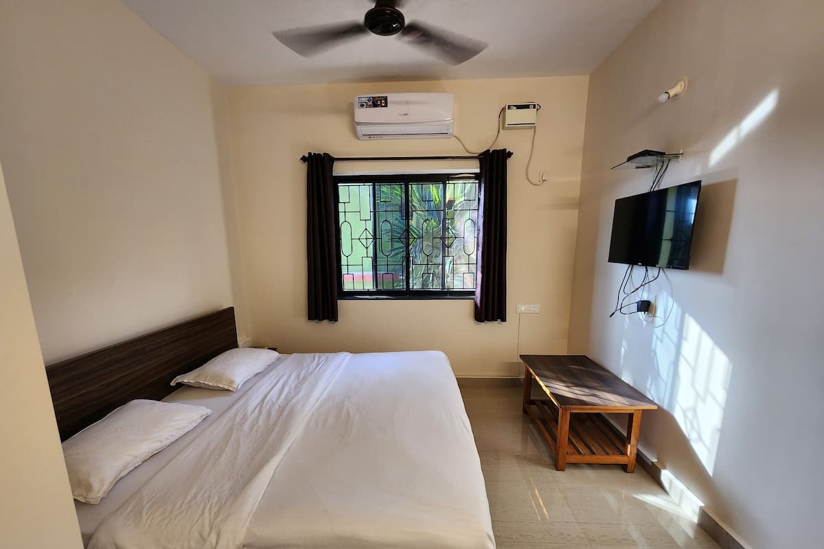 A hotel room with a bed, a ceiling fan, an air conditioning unit, a window with curtains, a TV mounted on the wall, and a small wooden bench.