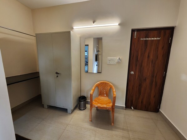 A room with a cabinet, a wooden door, a chair, a trash bin, and a mirror on the wall. - Sai Sharad Hotel (Pernem)