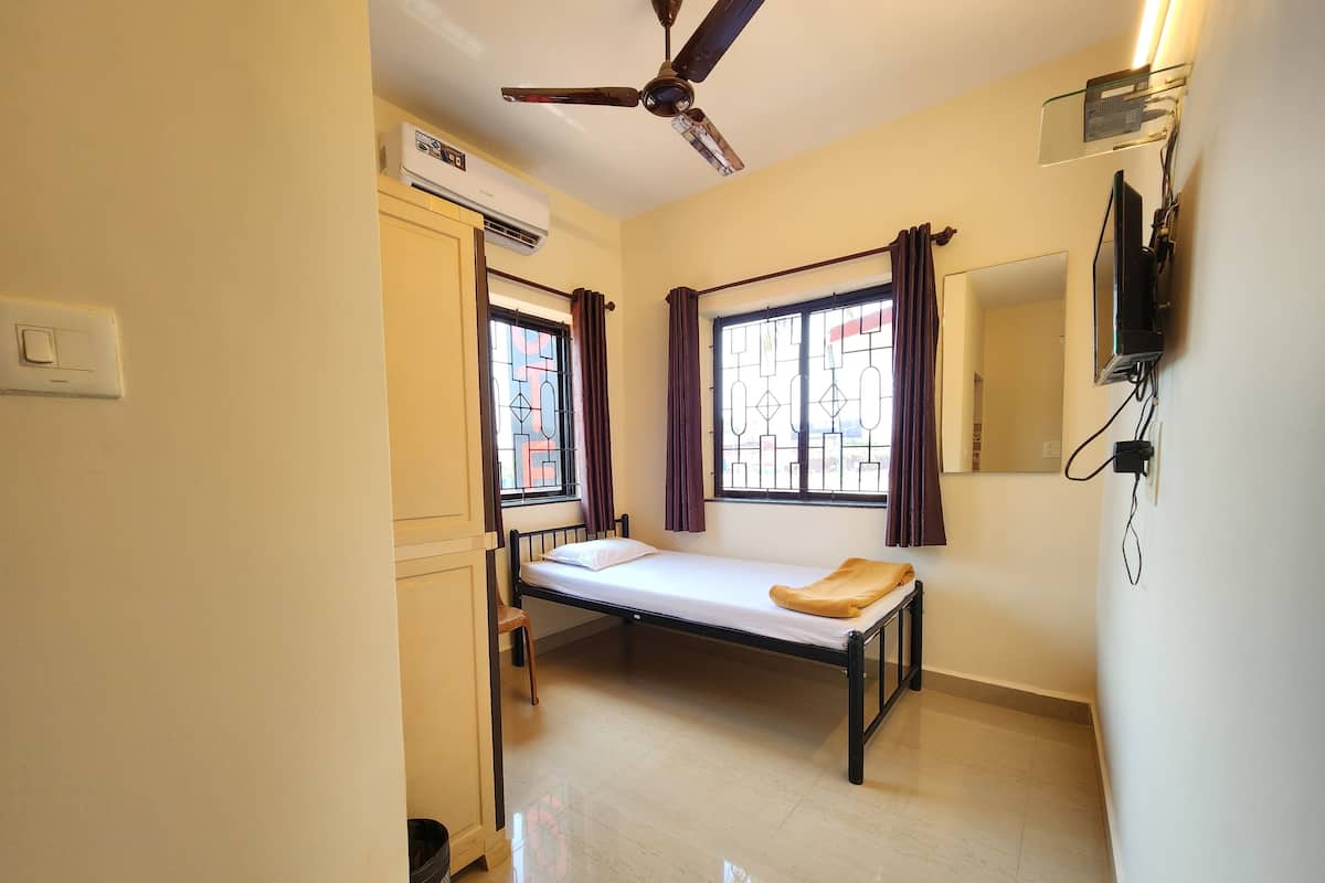 A small, clean room with a bed, a ceiling fan, an air conditioning unit, a window with curtains, and a wall-mounted TV.