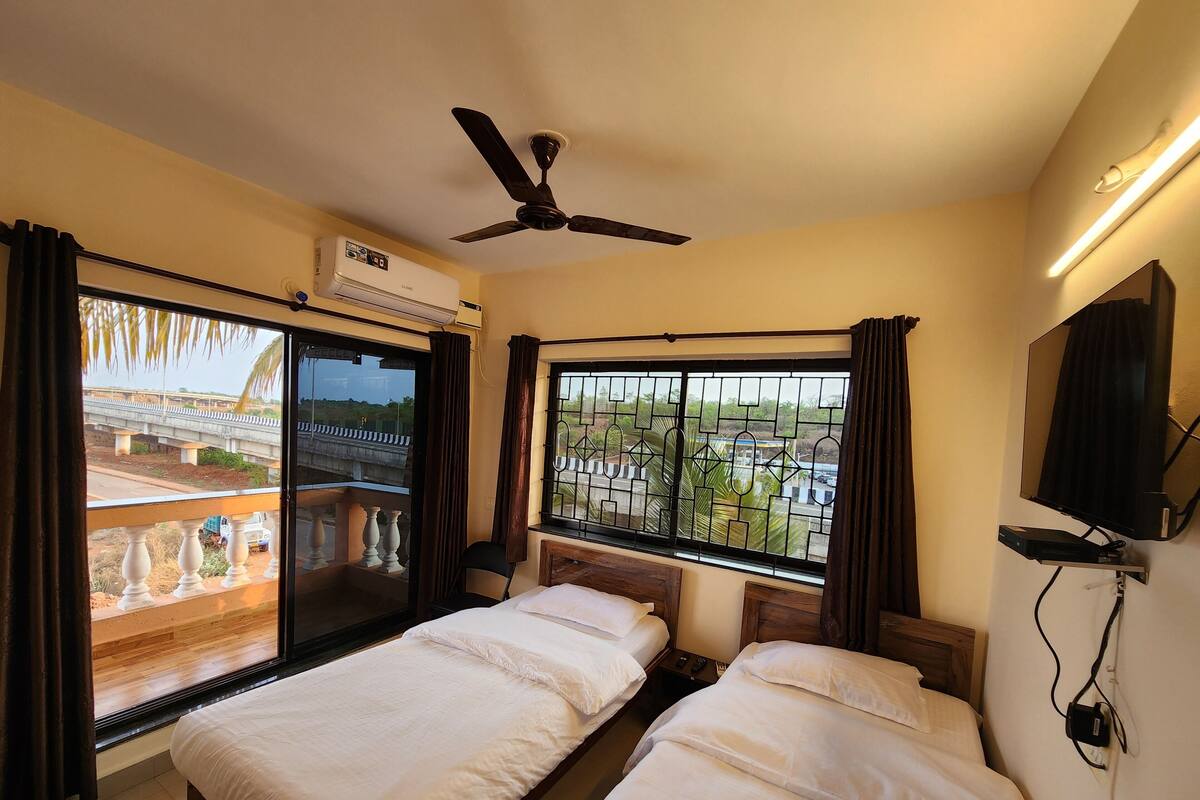 A hotel room with two beds, a ceiling fan, an air conditioning unit, a TV, and a window with a view of a bridge and greenery.