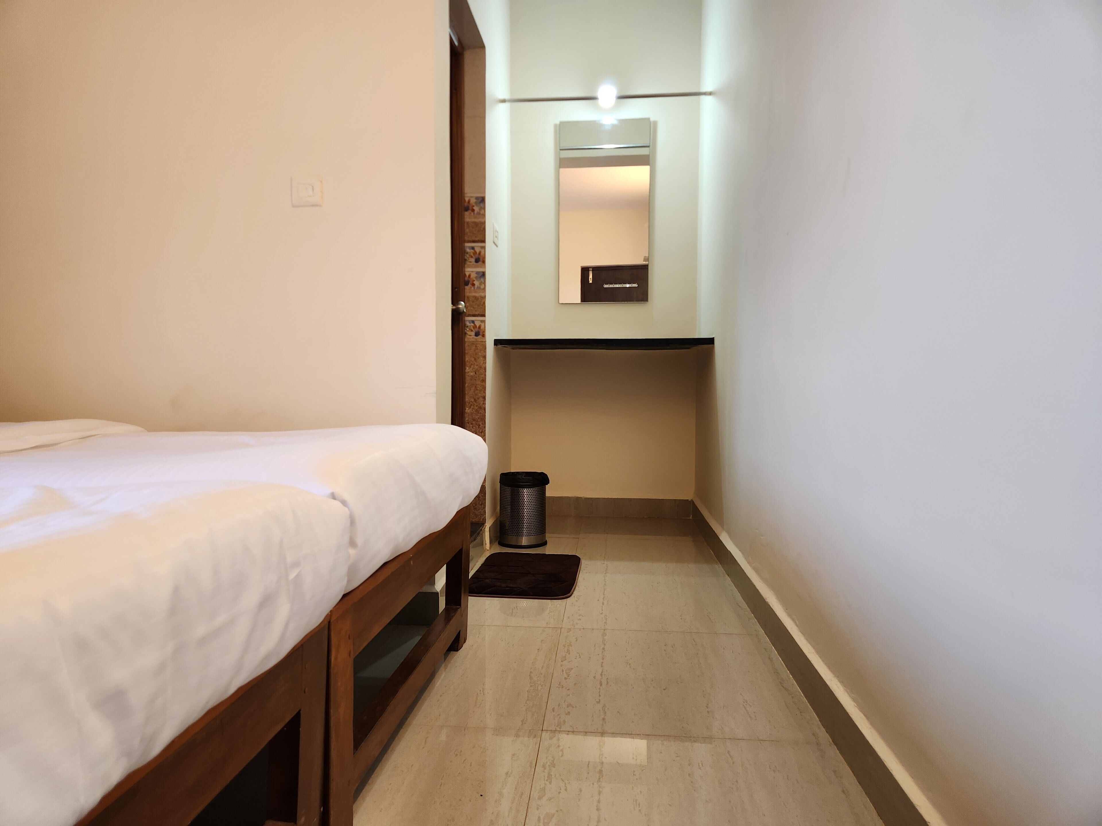 A narrow hotel room with a single bed, a small trash bin, and a mirror above a sink.