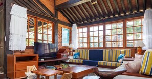 A cozy room with a wooden ceiling, large windows, a sofa with striped cushions, a television, and a wooden table.