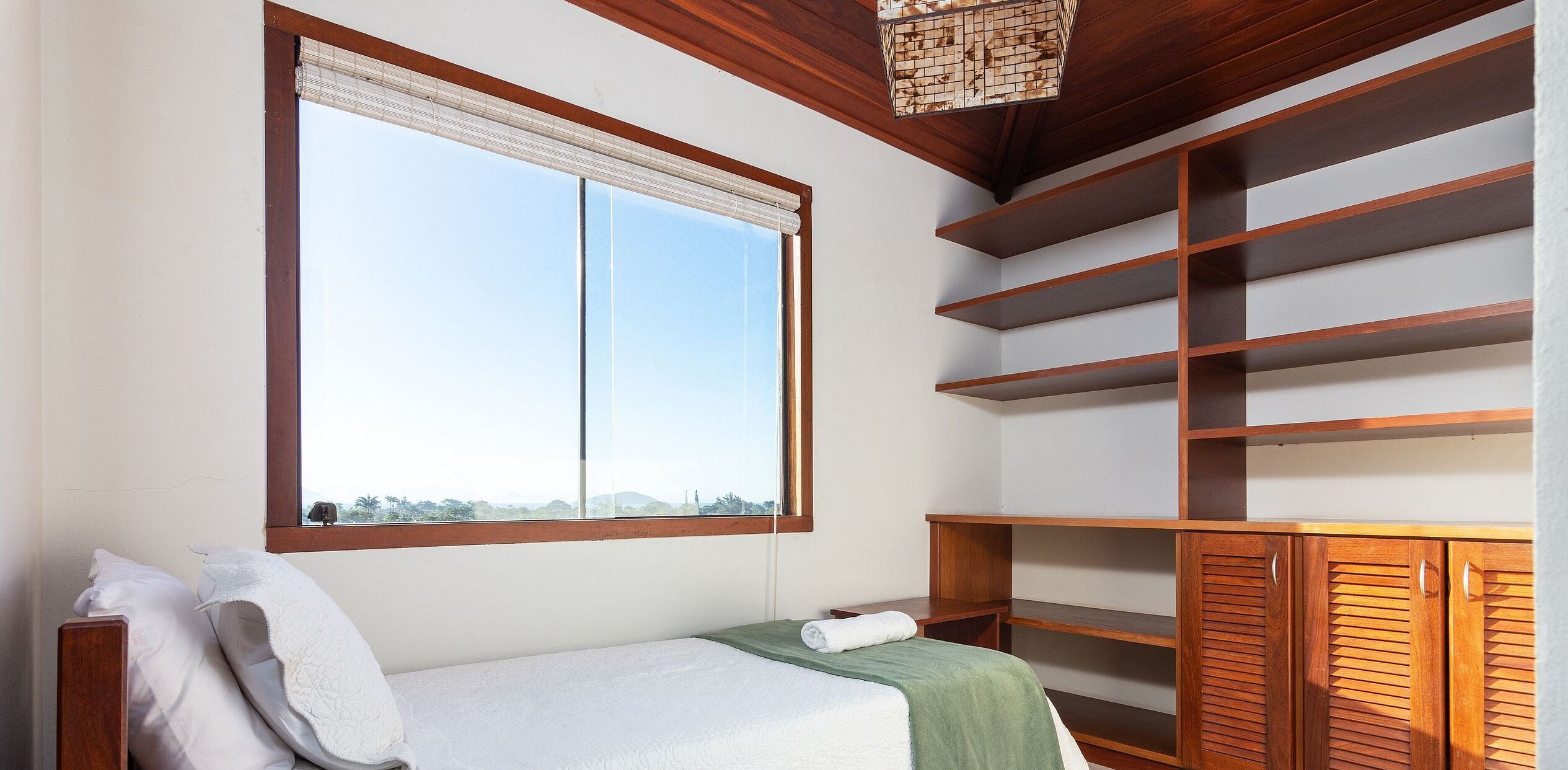 A room with a bed, a window with blinds, a wooden ceiling, and built-in shelves.