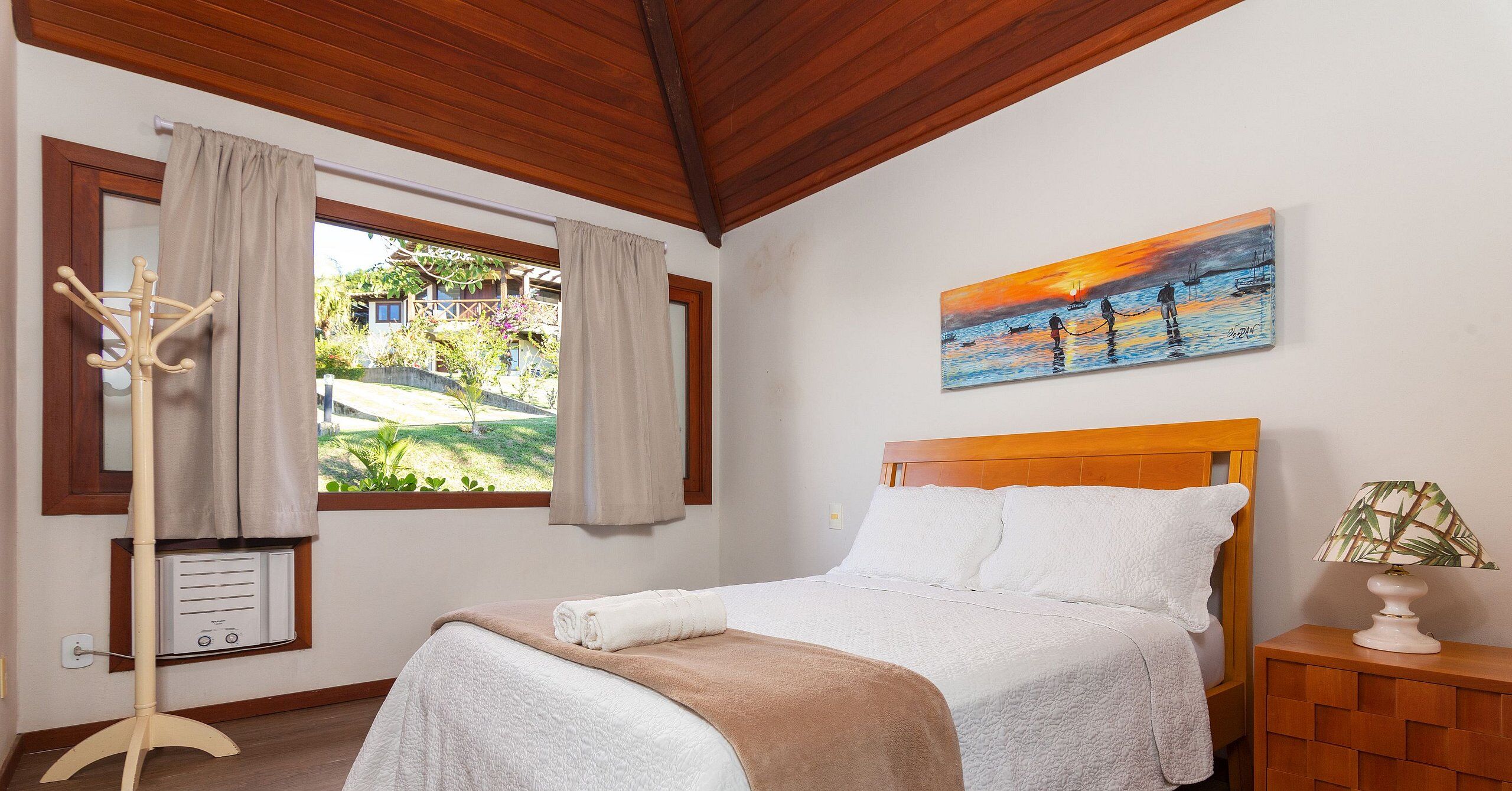 A bedroom with a wooden ceiling, a bed with white bedding, a painting of a sunset over water, and a window with curtains.