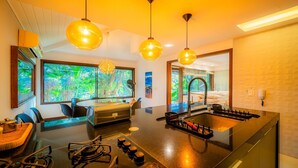 A modern kitchen with a central island, large windows with a tropical view, and pendant lighting.