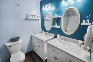 Une salle de bain avec deux lavabos, des toilettes et une douche. On y trouve des étagères avec des articles de toilette et un miroir au-dessus des lavabos.