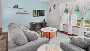 Ein Wohnzimmer mit grauem Sofa, einem Couchtisch, einem Fernseher und einem Fenster mit Vorhängen.