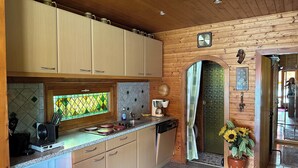 Une cuisine avec des meubles en bois, une fenêtre ornée d’un motif losange vert, une machine à café et une horloge au mur.