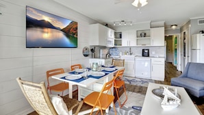 A modern kitchen with white cabinets, a dining area with orange chairs, a flat-screen TV, and a ceiling fan. - Charming beachfront villa in Hilton Head Island with pool, beach/pool bar & more (Hilton Head Island)