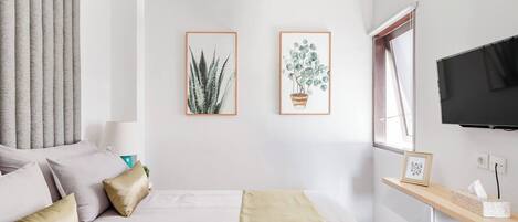 A modern bedroom with a bed, two framed plant artworks, a TV, and a bedside table with a lamp.
