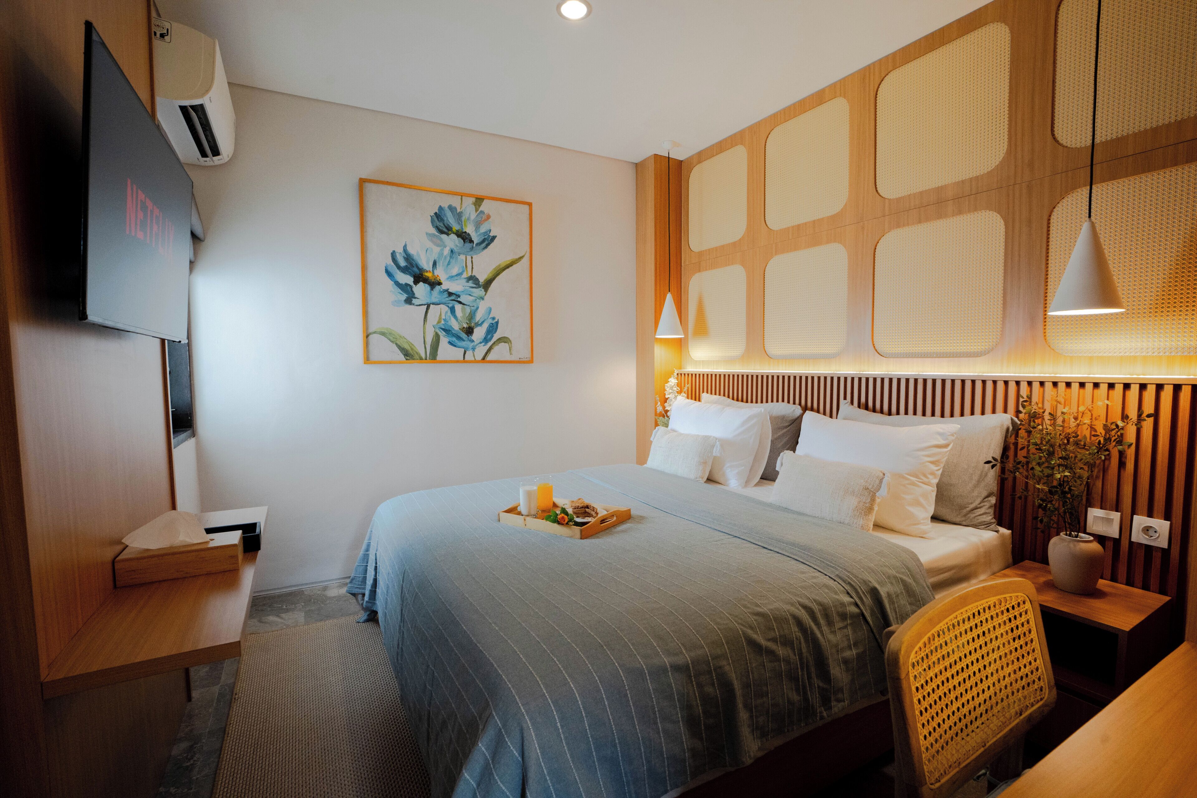 A hotel room with a bed, bedside table, lamp, artwork, and a TV.